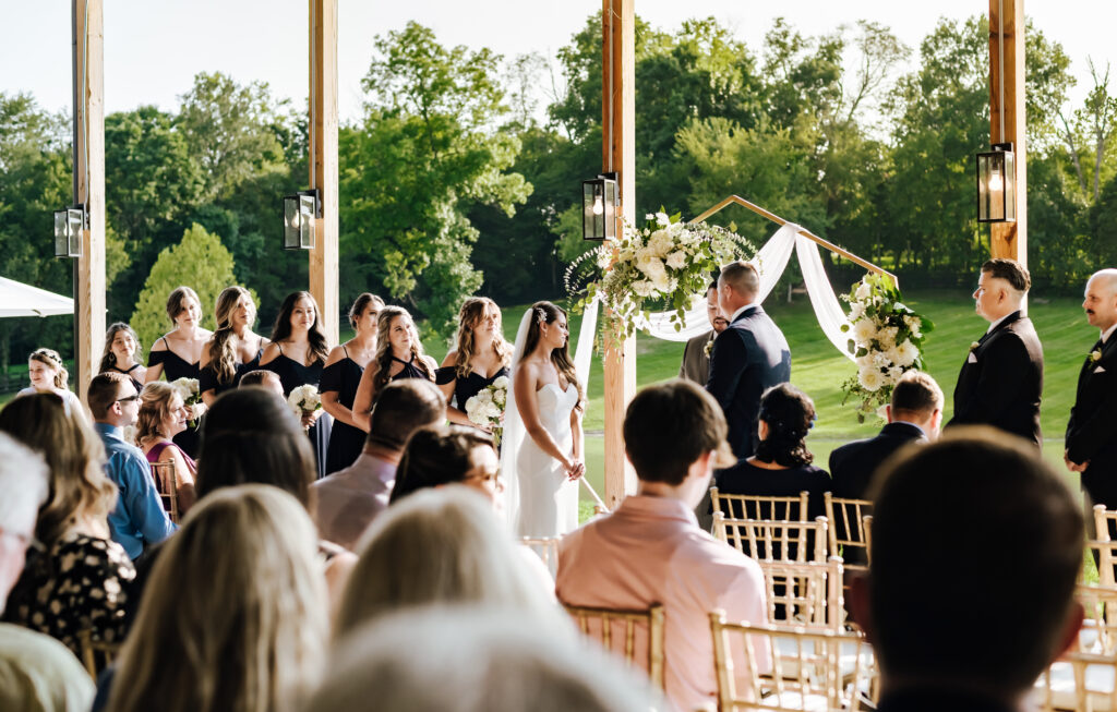 A bride and groom stand in front of a hexagon shaped arch with their wedding party. In the foreground, their guests look lovingly towards the couple