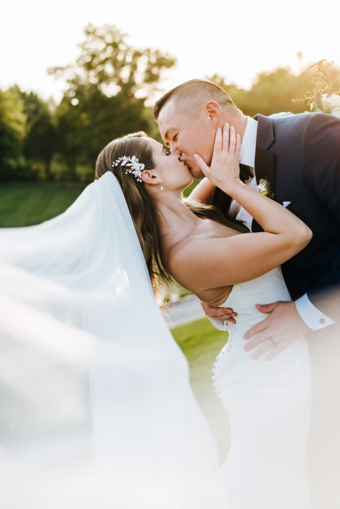 The groom dips the bride backwards for a romantic kiss. Her veil blows towards the camera and the glowy sun shines brightly behind them.