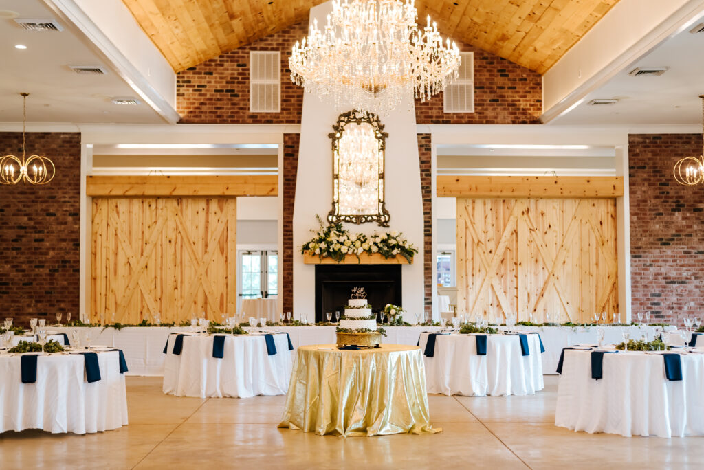 A wide photograph shows the grand ballroom at Hazelnut Farm. A large chandelier hangs in the center, in the back there is a fireplace with florals on the mantel, and a table with the wedding cake is in the center of the room, surrounded by round tables for guests.