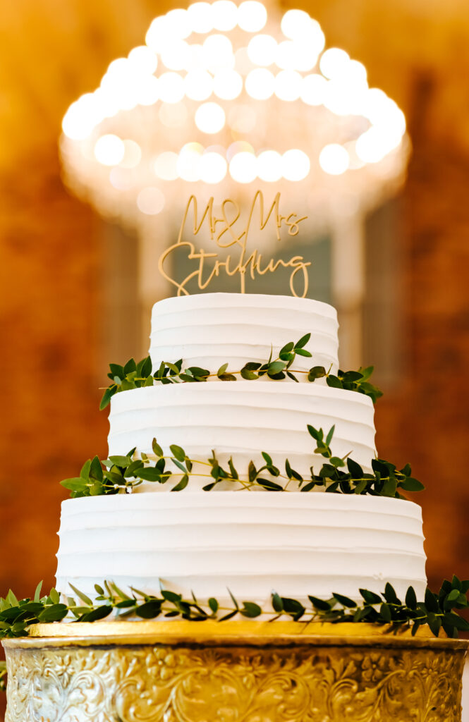 A three layer round cake with white frosting, greenery around the base of each layer, and a topper that says Mr. and Mrs. Stribling is elegantly placed atop a gold cake stand. In the background, you see the magnificent gold chandelier.