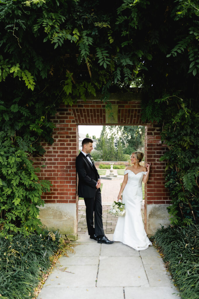 The bride and groom each lean against one side of a brick doorframe. Lush greenery covers the brick walls on either side.