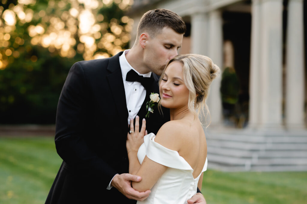 An elegantly dressed groom and bride embrace as the groom kisses the bride's forehead. They stand in front of the pillared staircase of Gardencourt.