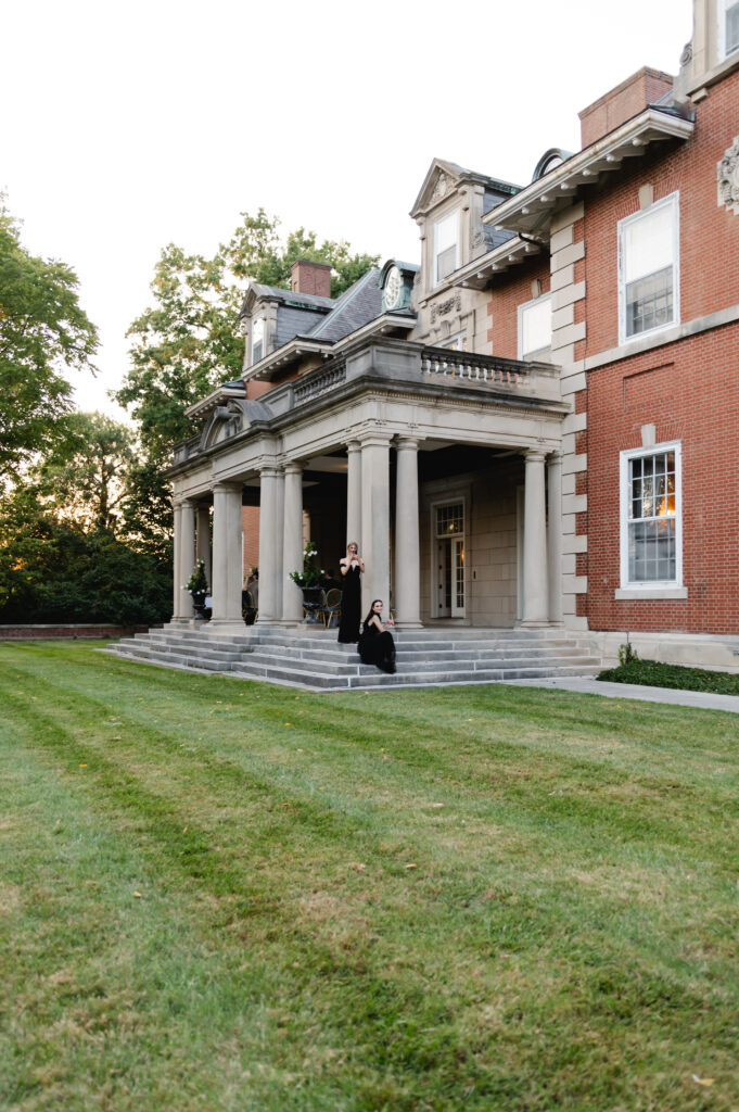 Two bridesmaids wearing full length black gowns sit on the steps of the pillared back entrance of Gardencourt