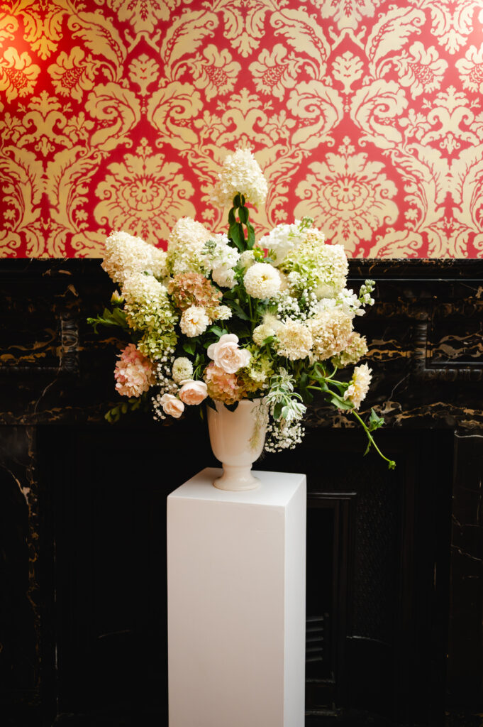 A large floral arrangement in a vase sits atop a rectangular pillar. The wall behind it features a vintage red wallpaper and dark wood.