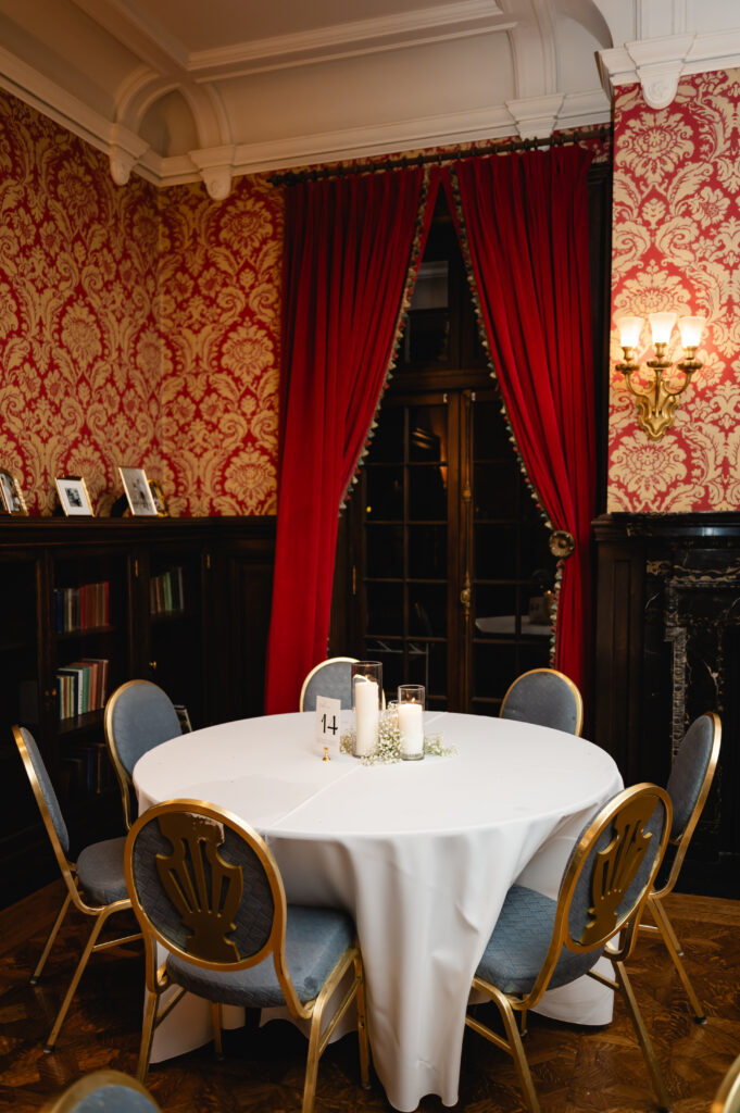 Another historic room within Gardencourt features red curtains and red vintage patterned wallpaper. There is a guest table with French blue chairs, pillar candles and florals.