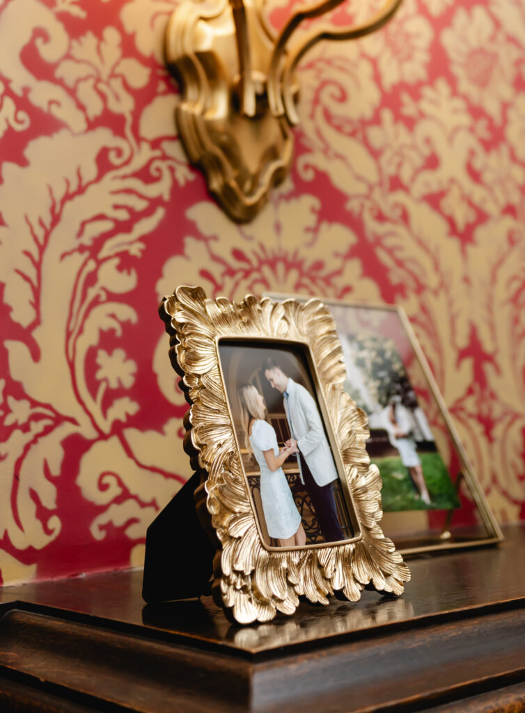 A golden frame containing an engagement photo of the couple sits on a mantel in front of red vintage wallpaper