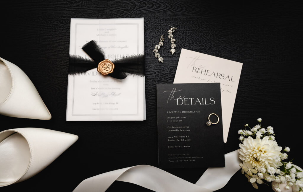 A black and white invitation suite is set up in a flatlay arrangement with jewelry, flowers and florals around the invitations