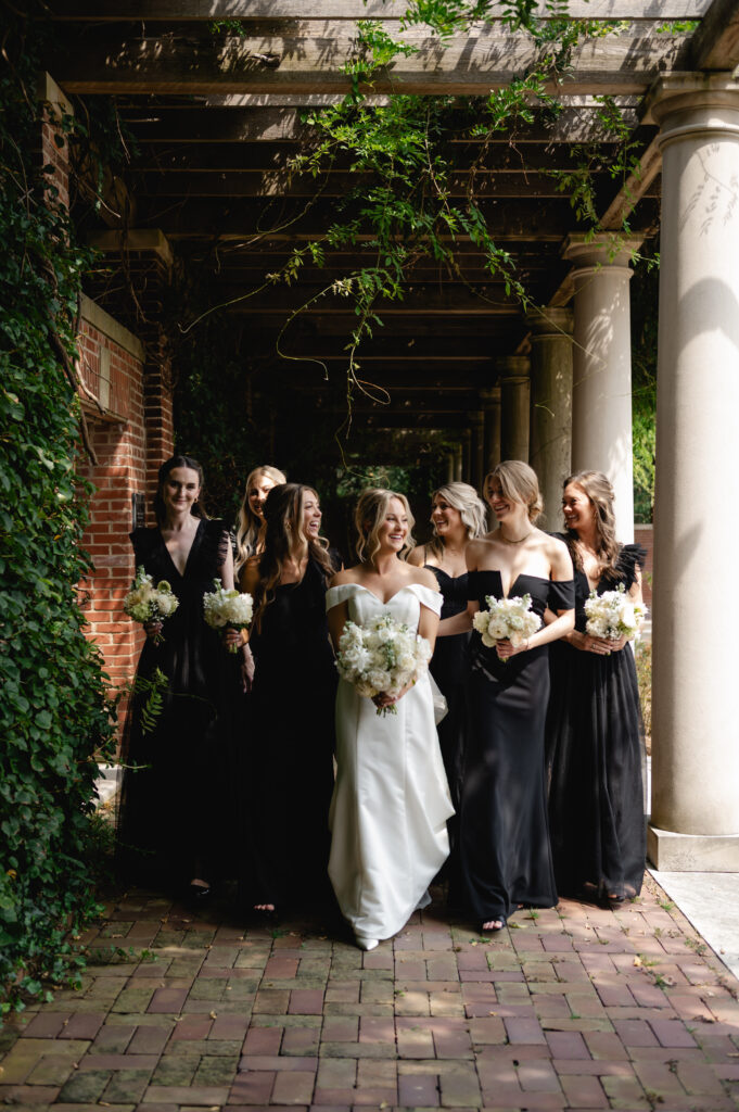 The bridesmaids in full length black gowns look sweetly at the bride. All hold their white and green bouquets as they walk through the colonnade.