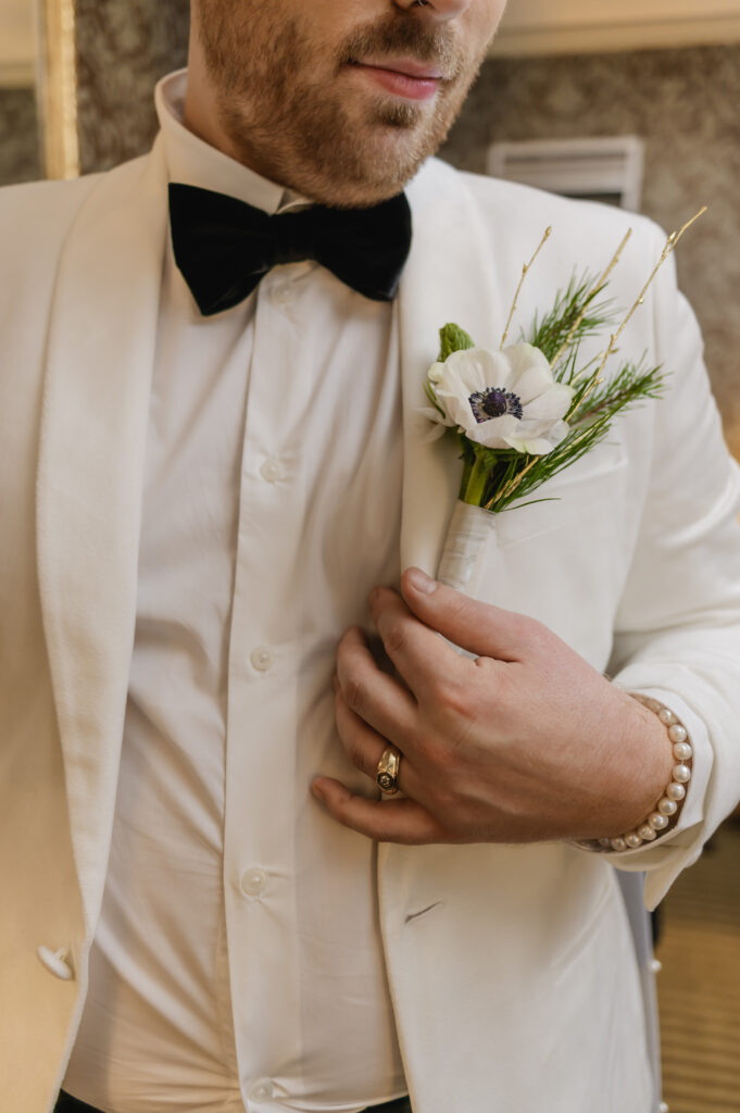 A close up shows the anemone boutonniere pinned on the groom's white tux 