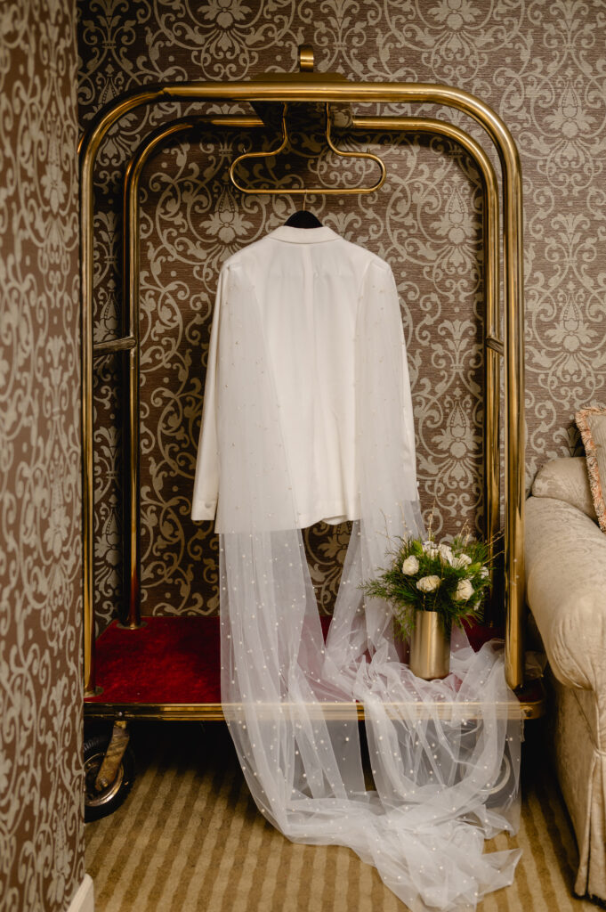 A hotel cart showcases one of the grooms' white tuxedo jacket with two pearled cape attachments (one for each shoulder). A golden vase with a bouquet sits stylishly on the cart.