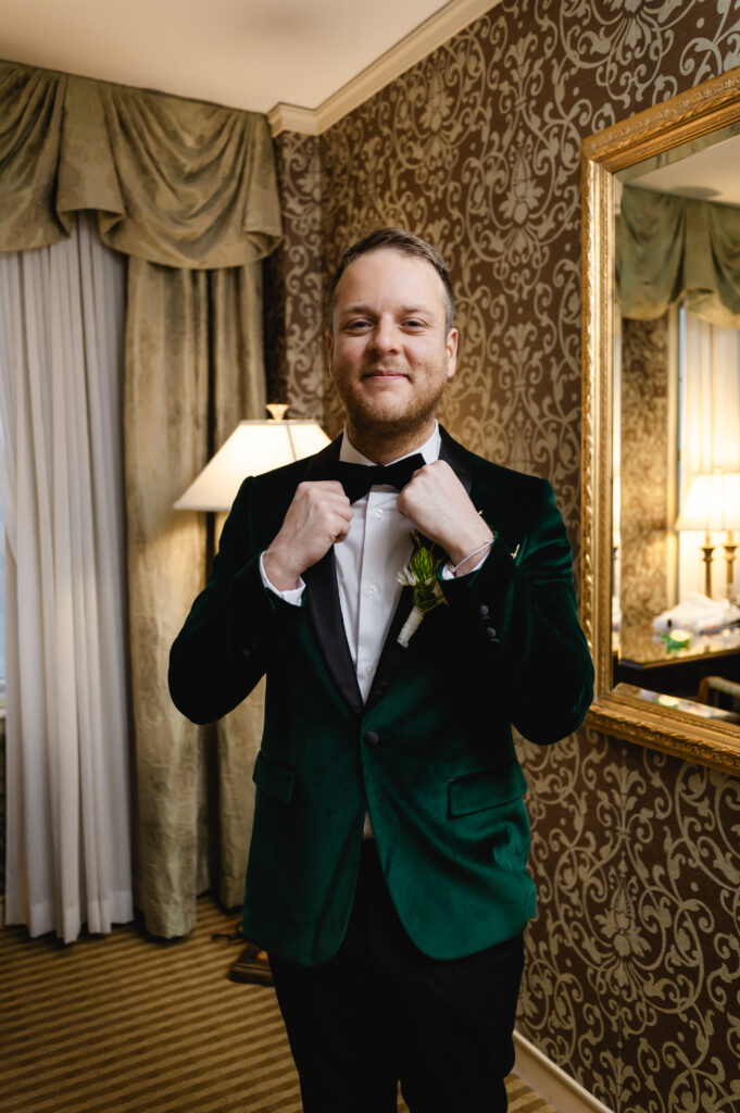 The smiling groom adjusts his bowtie. He wears a velvet emerald tux jacket with a white dress shirt and black pants.