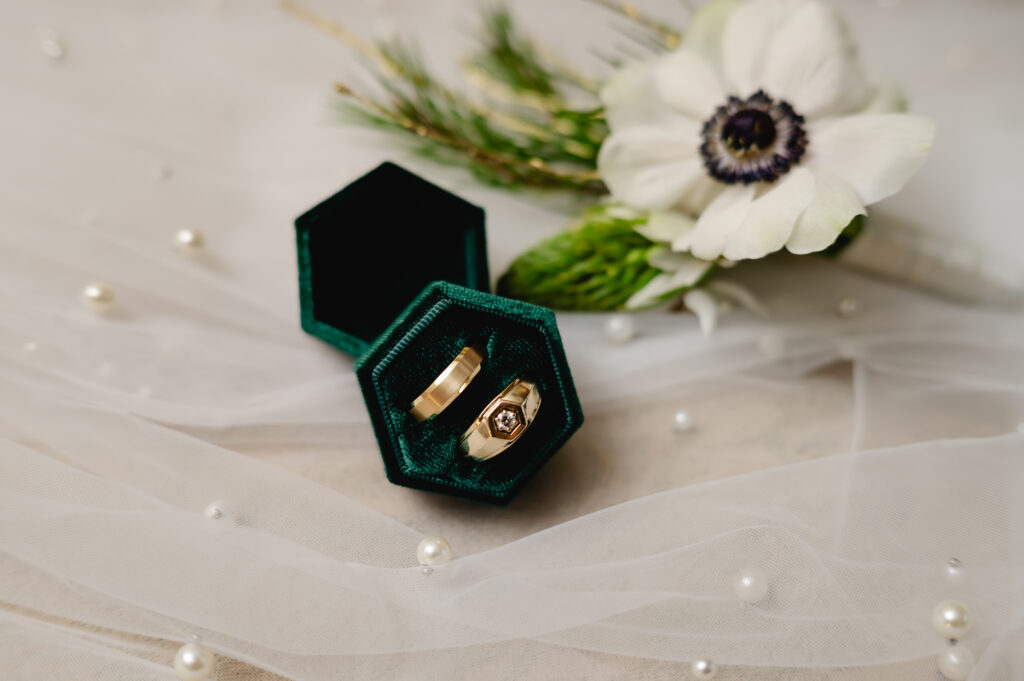 An emerald green ring box holds two gold bands. It sits atop a pearled veil and next to it is a boutonniere with a large anemone flower.