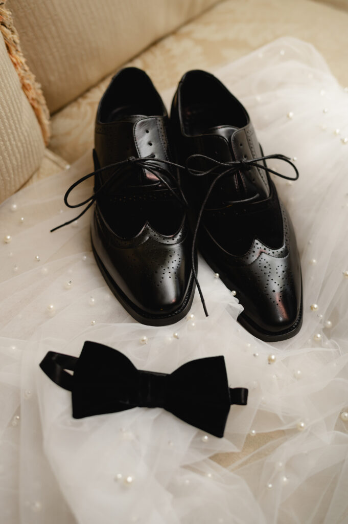 A groom's shoes and bowtie lay askance on a pearled white veil