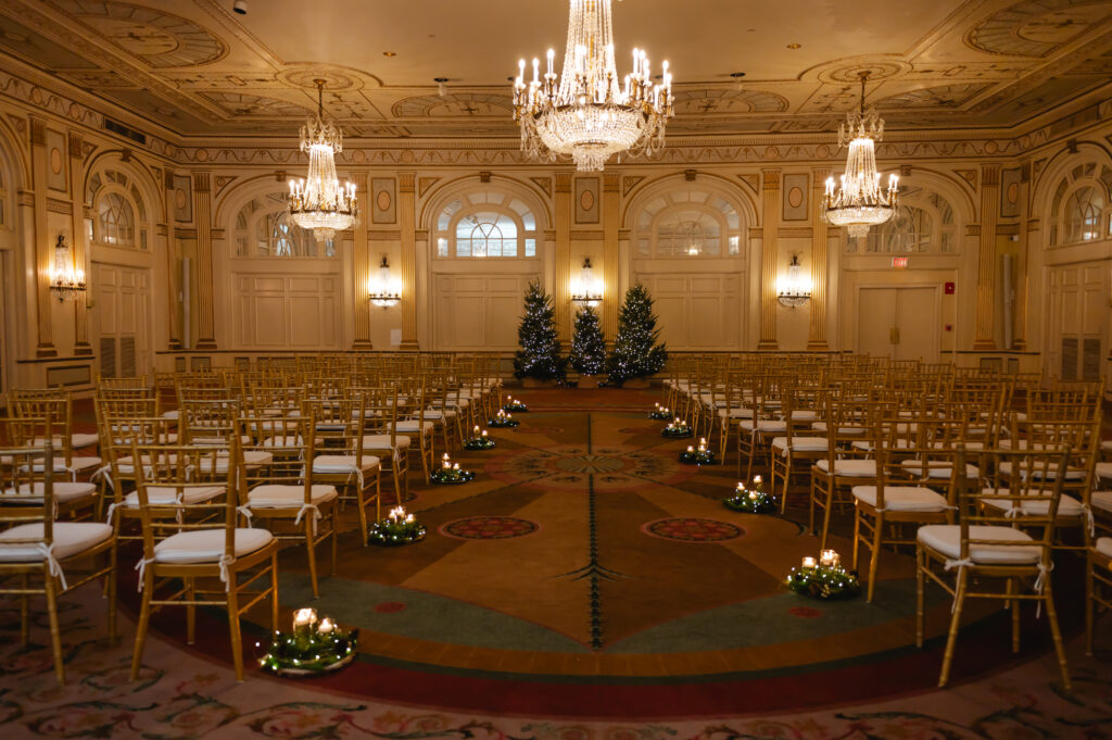 A landscape photos shows a stunning ballroom at the historic Brown Hotel. There are several ornate chandeliers, three Christmas trees in the center, gold chiavari chairs for guests and pillar candle centerpieces at the end of each aisle.