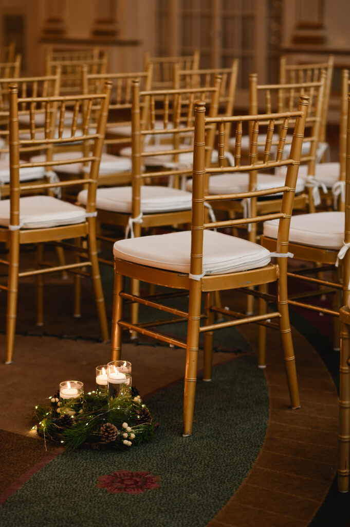 Gold chiavari chairs are set up for ceremony. Next to them in the aisle are three different height pillar candles with winter greenery around them