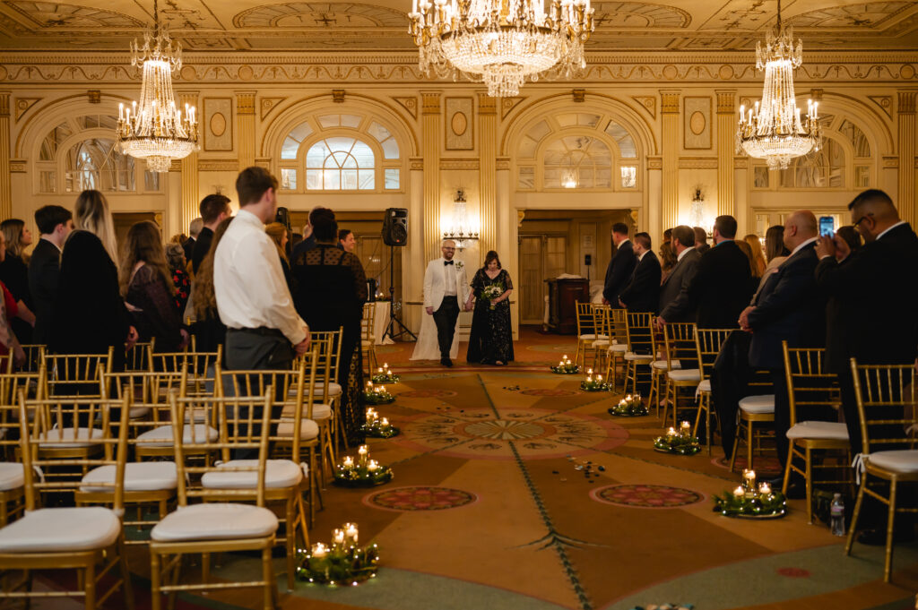 Groom Jared enters the ballroom escorted by his partner's mother, as everyone looks lovingly towards them