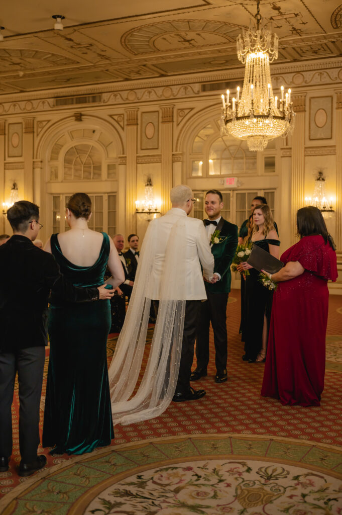 The grooms hold hands and their wedding party looks towards them