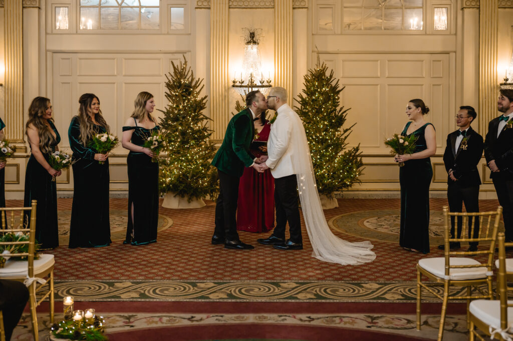 The grooms share a kiss as their wedding party smiles and cheers 