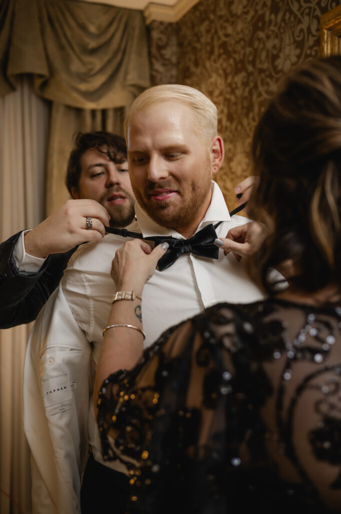 The groom's best man and mother in law help him put on his tux jacket and bowtie