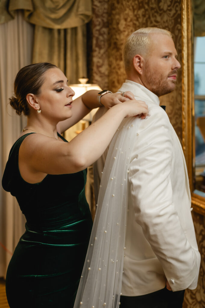 The maid of honor helps pin the pearled veil cape onto the shoulder of the groom's white tux jacket