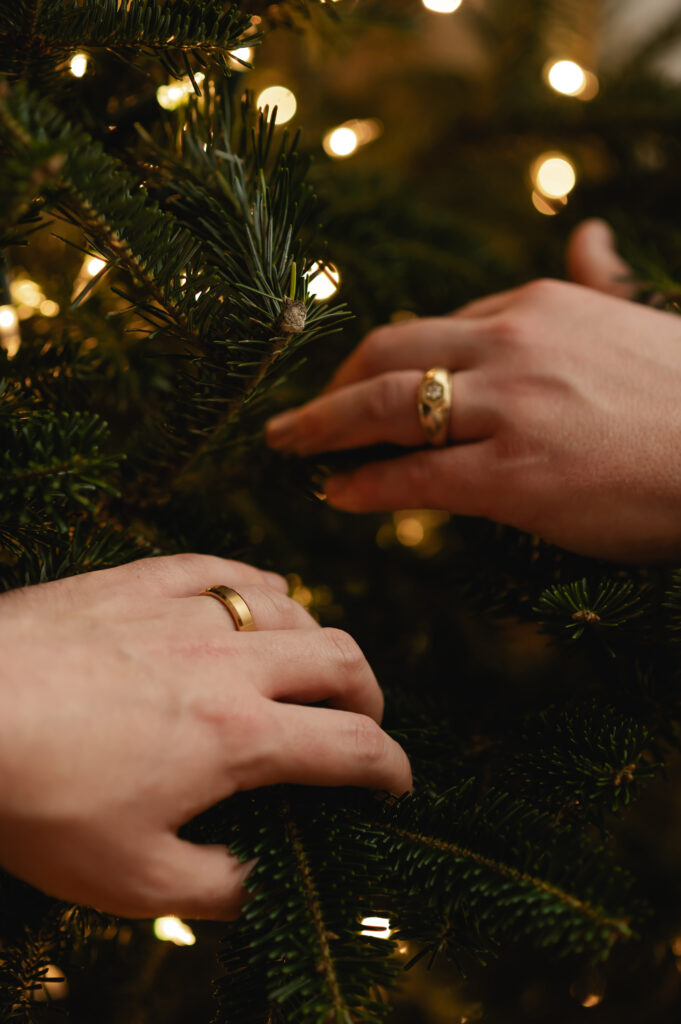 The grooms each hold the hand with their wedding band in front of the lit Christmas tree 