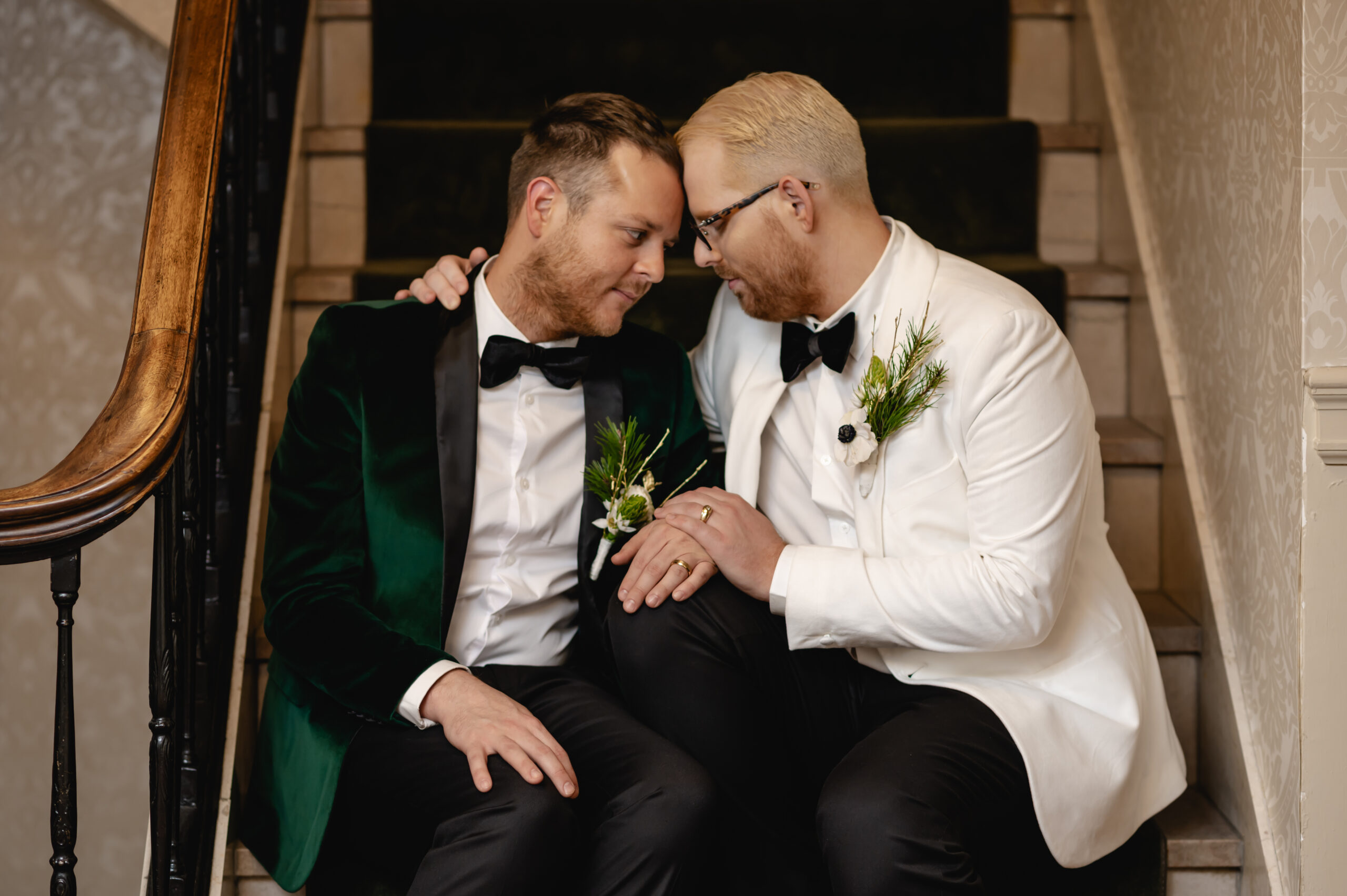 Two grooms sit on the staircase of the historic Brown Hotel and hold hands as they gently rest their foreheads together