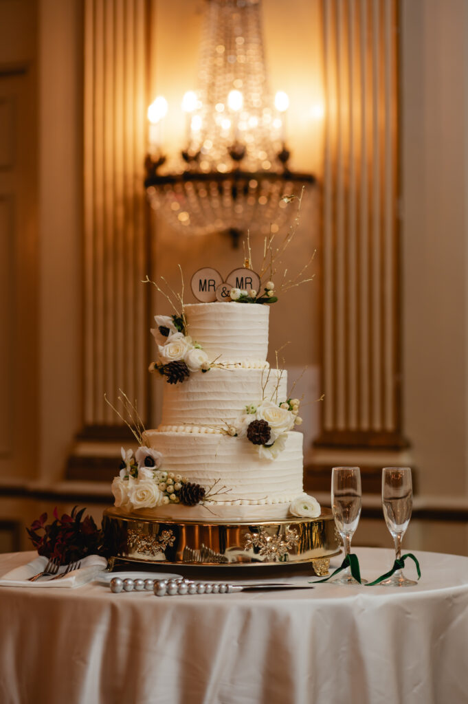 The cake table has a three tier cake atop an elegant gold base. Two champagne glasses sit next to the cake, and a small sconce chandelier glows behind the table.