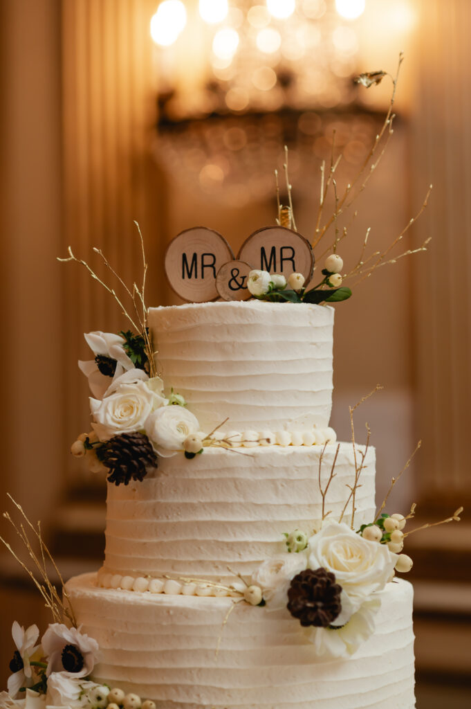 A three tiered round cake with pear accents at the base layers has "Mr. and Mr." toppers engraved in two small pieces of wood. Small wintry floral accents adorn the cake.