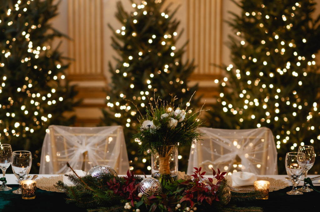 A sweetheart table decorated with winter greenery and small disco balls sits in front of three Christmas trees.