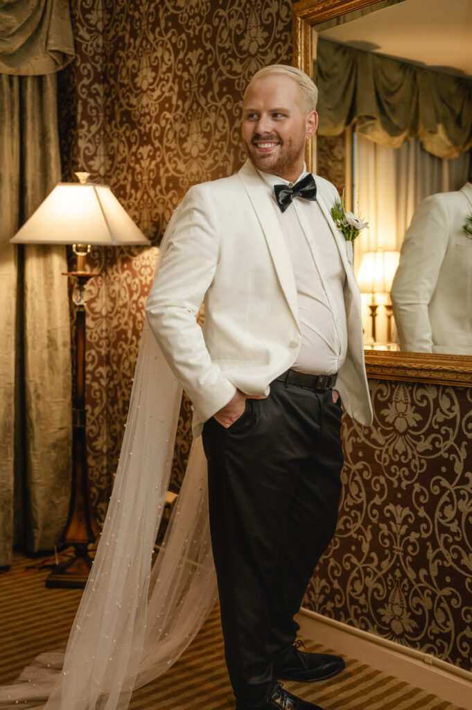 The groom smiles as he looks over his shoulder while wearing his white tux jacket, bowtie and black pants. A pearled veil cape drapes from both shoulders.