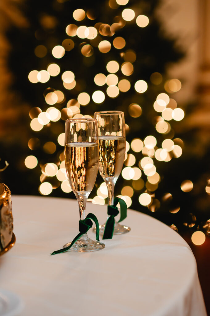 Two filled champagne glasses with emerald ribbons around the base are placed in front of two Christmas trees whose lights are softly blurred in the background