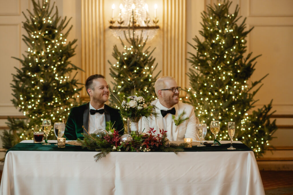 Matthew and Jared sit at their sweetheart table. Three lit Christmas trees are behind them, and they smile as they listen to their friends and family share speeches