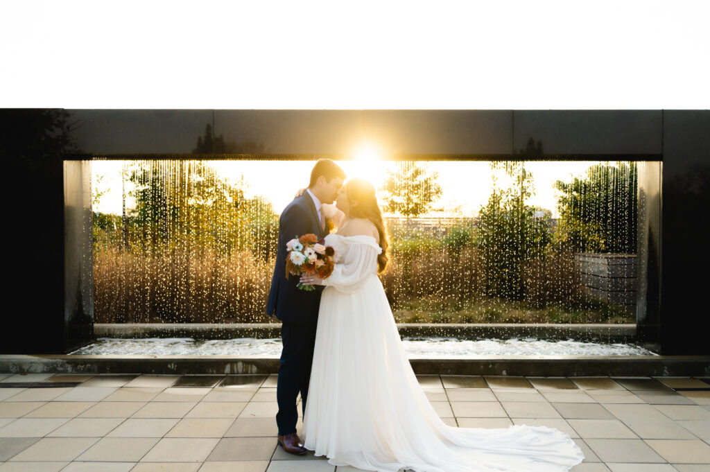 A couple embraces in front of the modern fountain sculpture as the sun beams brightly behind them