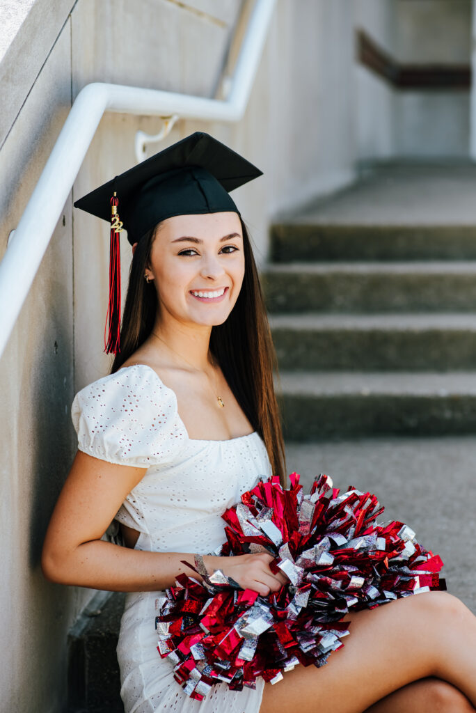 A cheerleader sits on the steps outside her school wearing a white dress, black graduation cap, and holds her red, black, and white pom poms in her lap