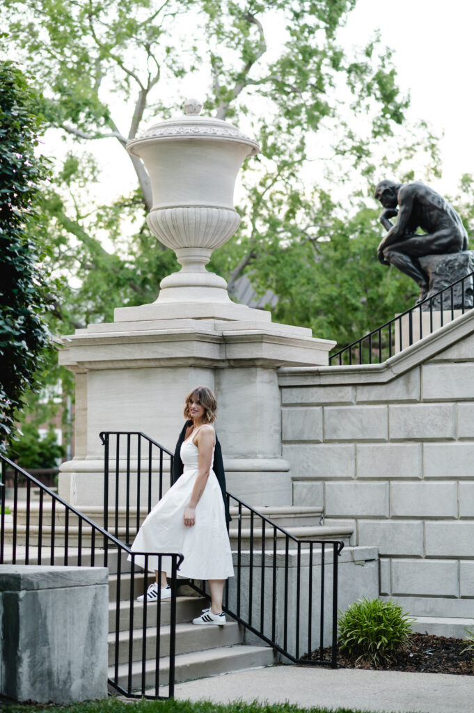 A University of Louisville graduate walks up the stone staircase in a beautiful scene of lush green trees. The bronze Thinker Statue is in the background.