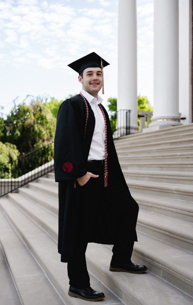 A male grad wearing black slacks, robe and cap with a white dress shirt stands on the steps of his school campus