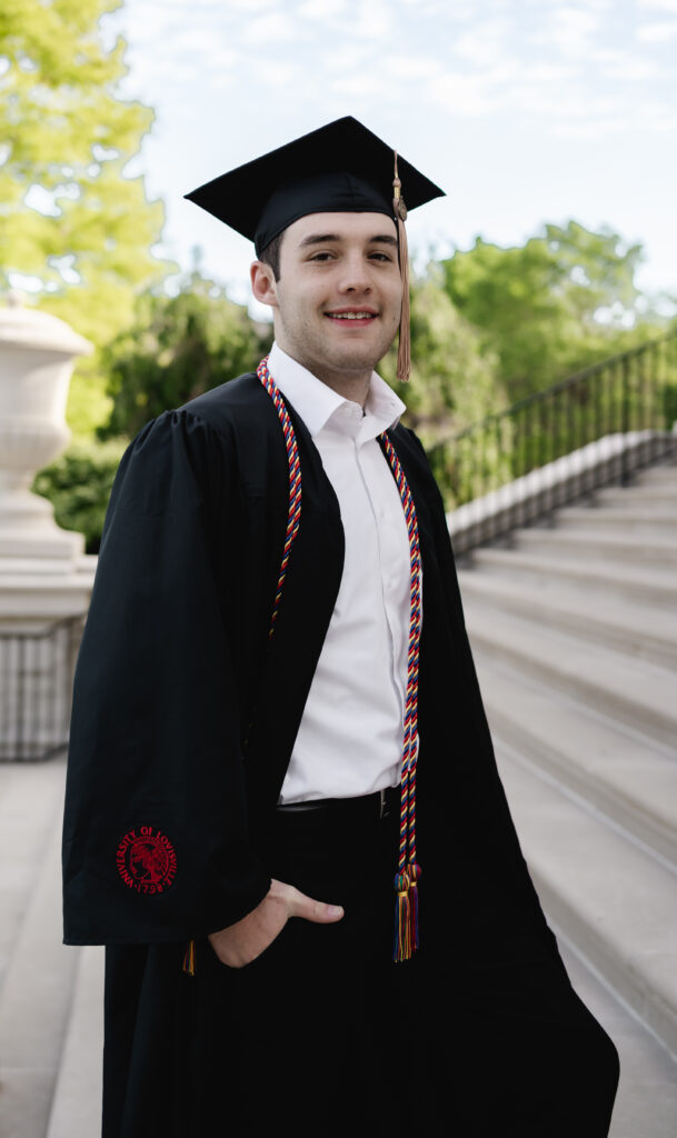 A male UofL grad strikes a cool pose with his hand in his pocket. He wears the black graduation attire and a white dress shirt.