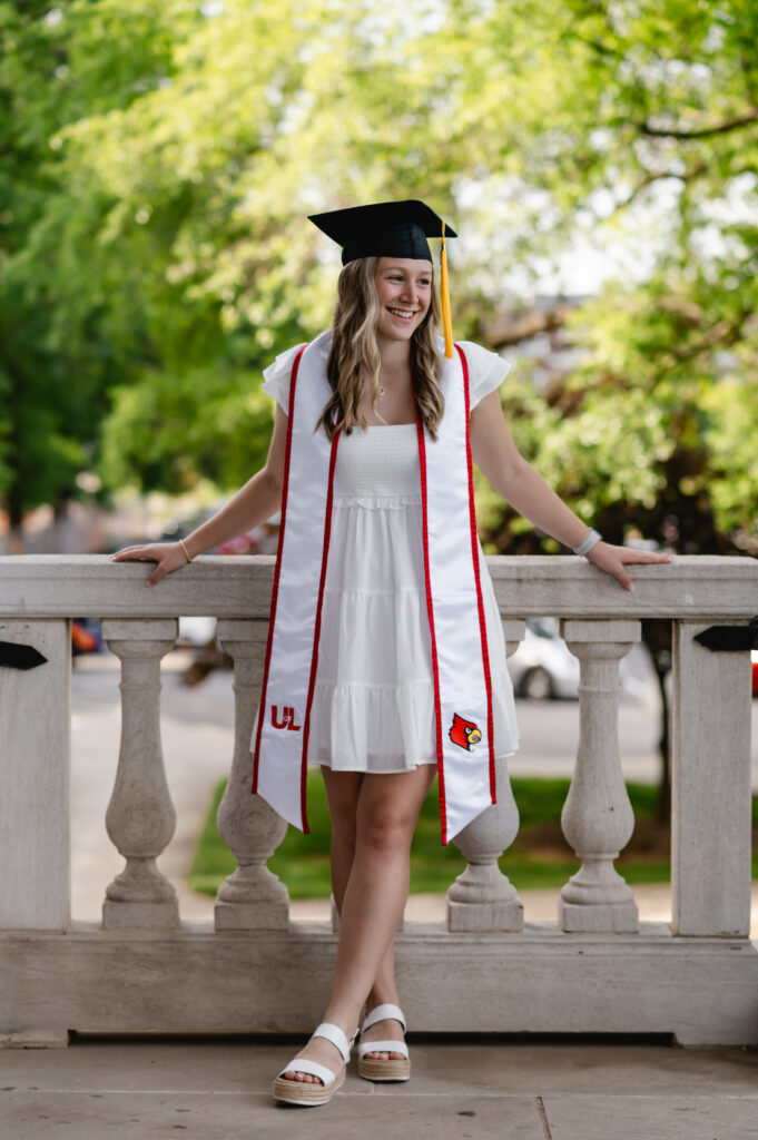 A UofL grad in a white gown, graduation cap, white stole, smiles as she poses in front of a stone balcony with lush greenery behind her