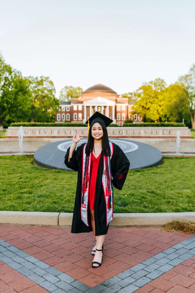 A University of Louisville graduate wearing a red dress, black heels and her graduation attire holds up the "L" symbol for her school in front of the impressive law school building