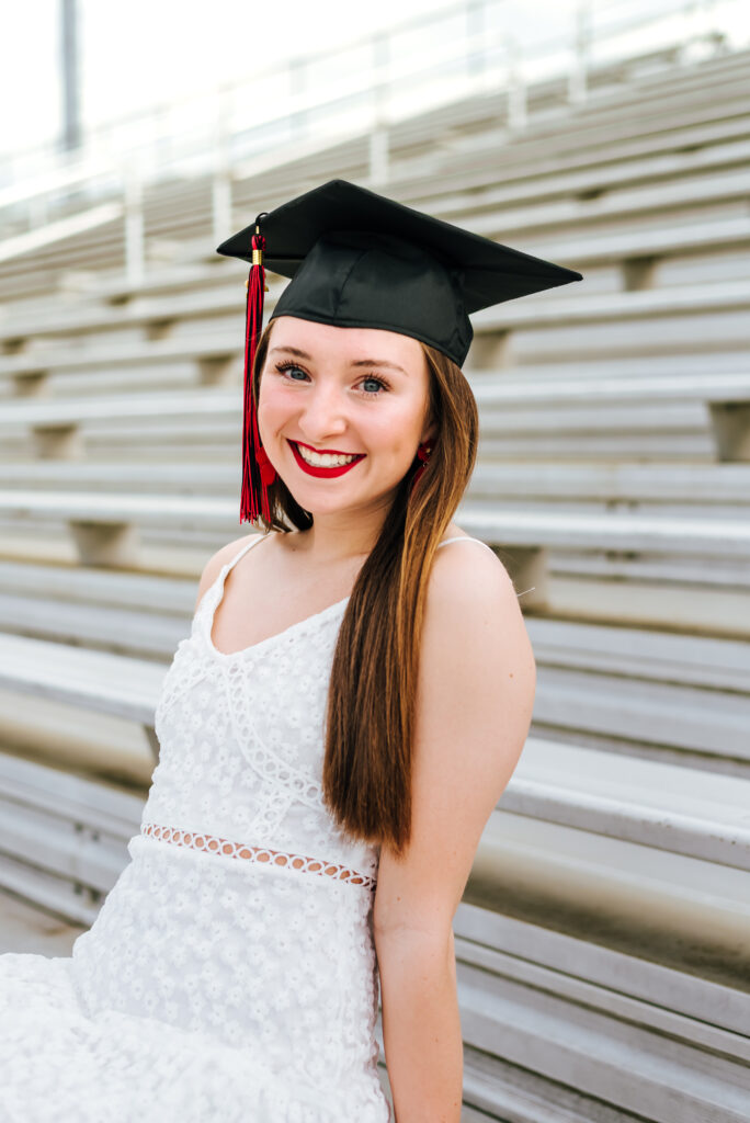 A grad in a white dress smiles radiantly as she sits in the bleachers of her school stadium. She wears bold red lipstick to match her school colors and her black grad cap and tassel.