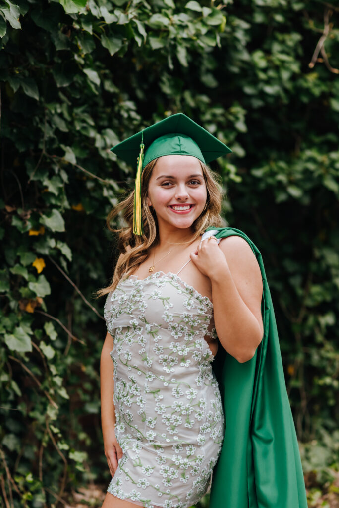 A high school grad wearing a floral spring dress holds her kelly green gown over her shoulder and wears her graduation cap