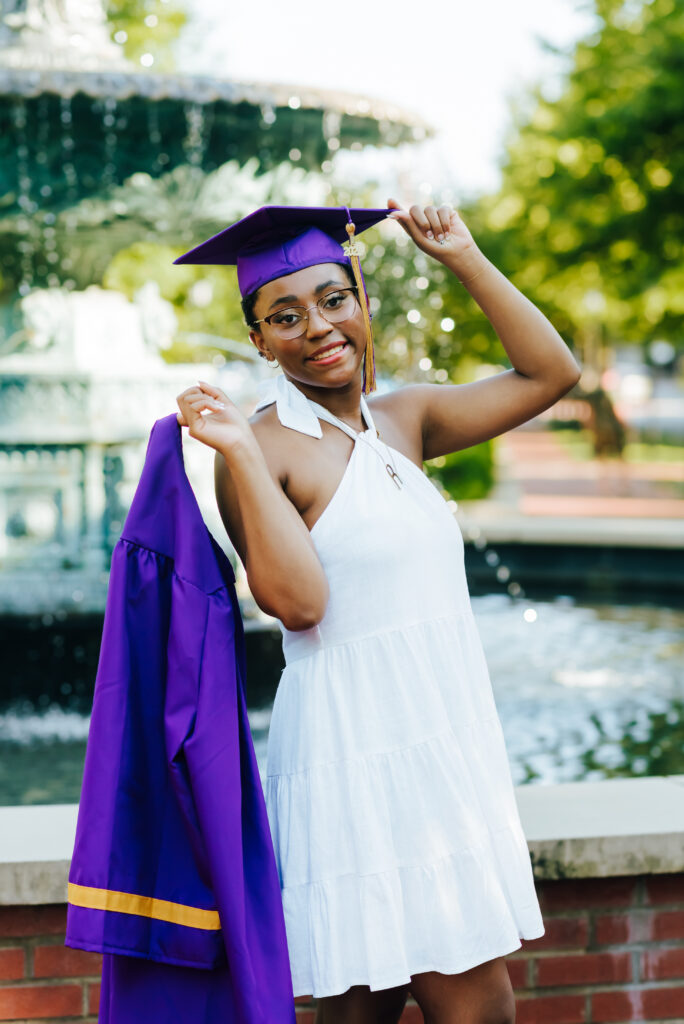 A high school graduate wearing a white dress drapes her purple graduation gown over her shoulder and holds her purple cap