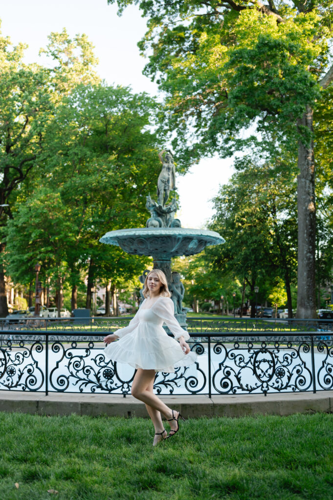 A high school grad in a white dress twirls in her dress in front of a beautiful fountain
