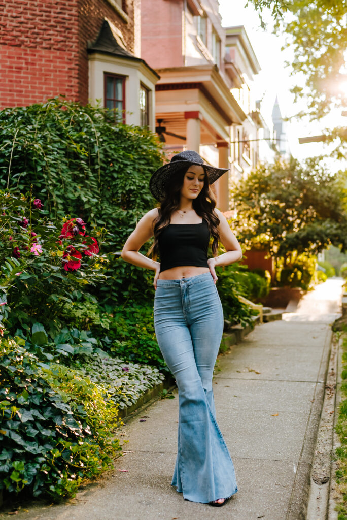 A stylish high school grad in jeans, a black top, and black hat poses with her hands on her hips in the historic St. James Court.