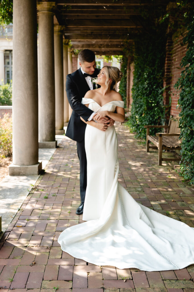 An elegantly dressed groom and bride embrace as the groom kisses the bride's forehead. They stand in front of the pillared staircase of Gardencourt.