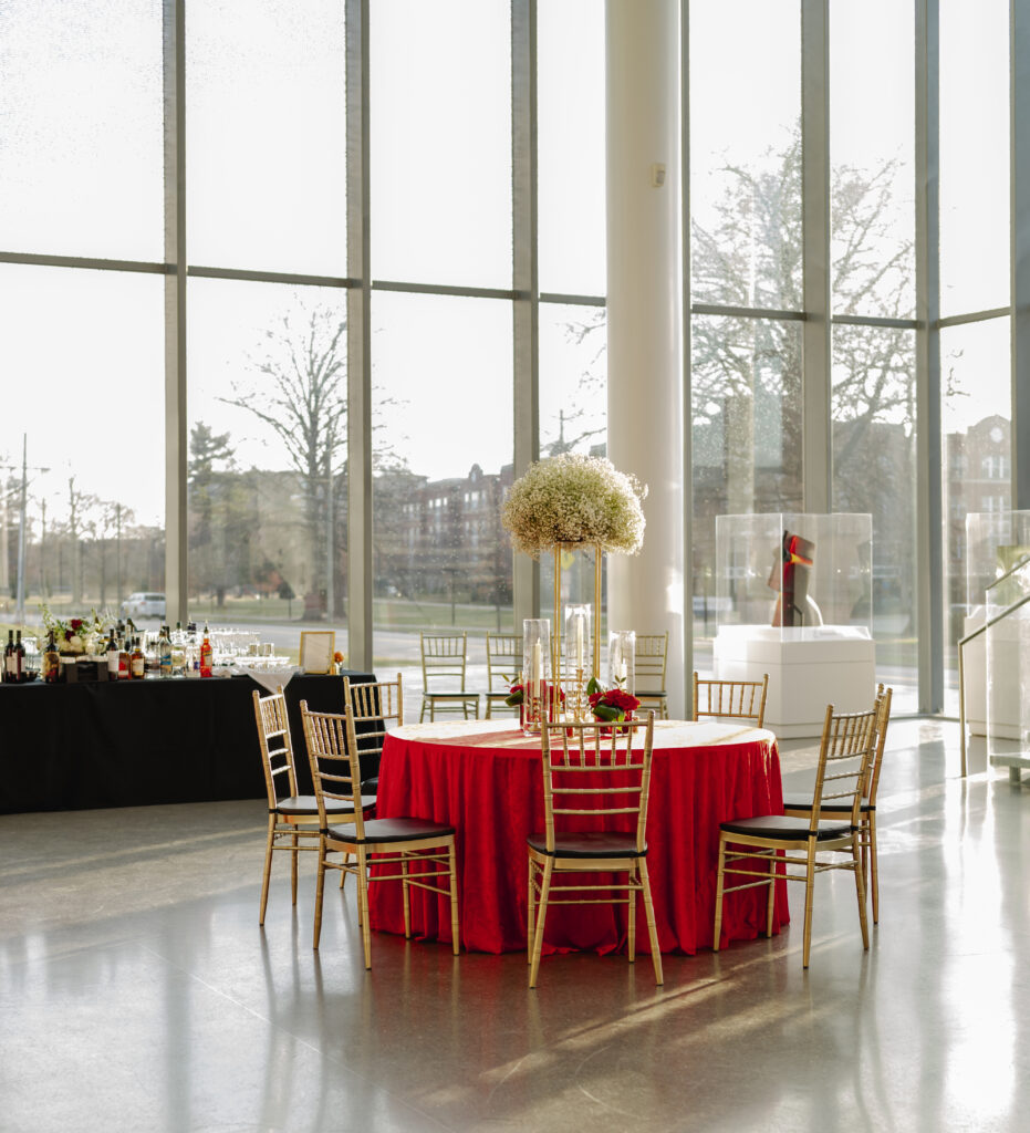 The atrium of the Speed Art Museum is decorated for cocktail hour with high top tables and red tablecloths, tall floral centerpieces and a bar.
