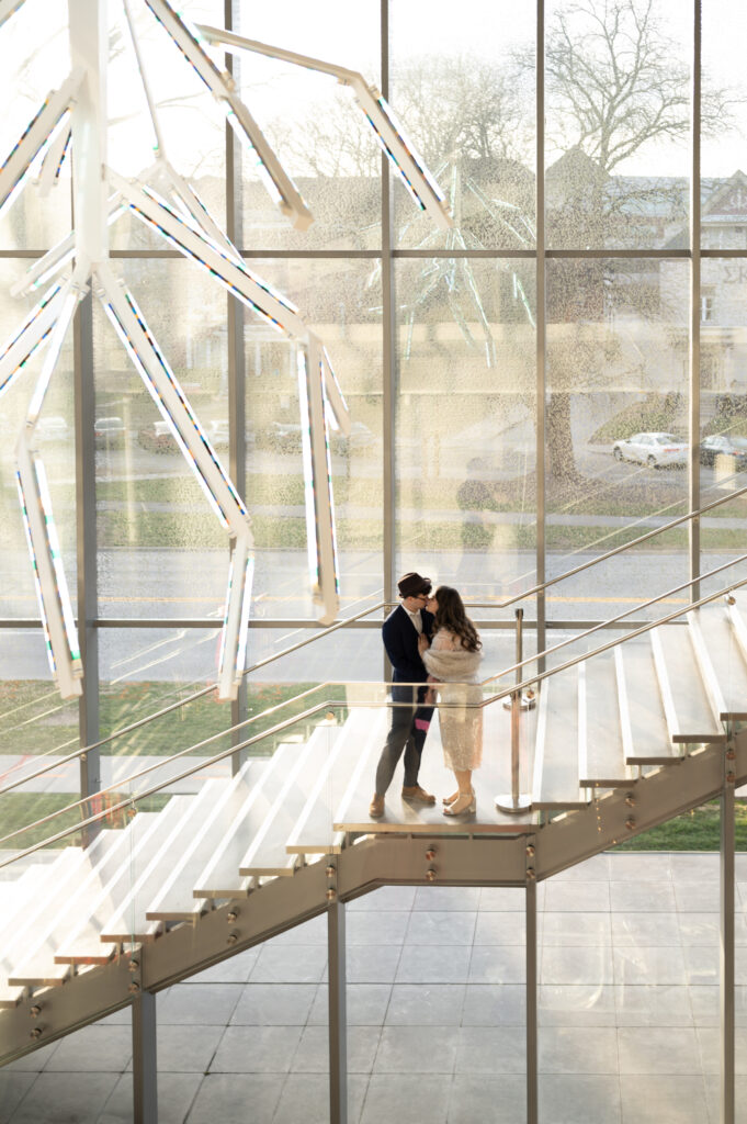 An impressive view from the staircase of the Speed Art Museum shows the floor to ceiling windows, decorative chandelier and a couple embracing.