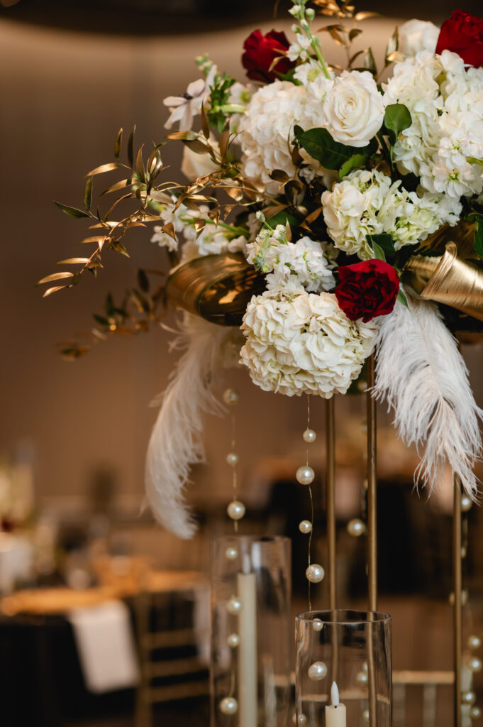 A closeup photo of an elegant centerpiece, with white and red roses, greenery, feathers, and pearls.