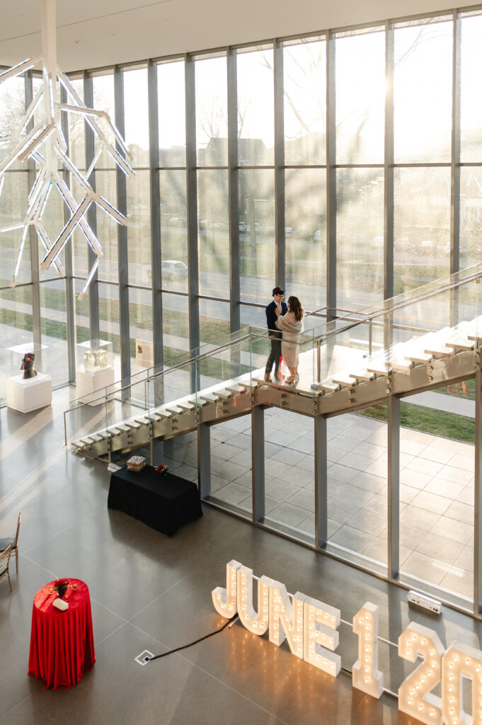 An impressive view from the staircase of the Speed Art Museum shows the floor to ceiling windows, decorative chandelier and a couple embracing.