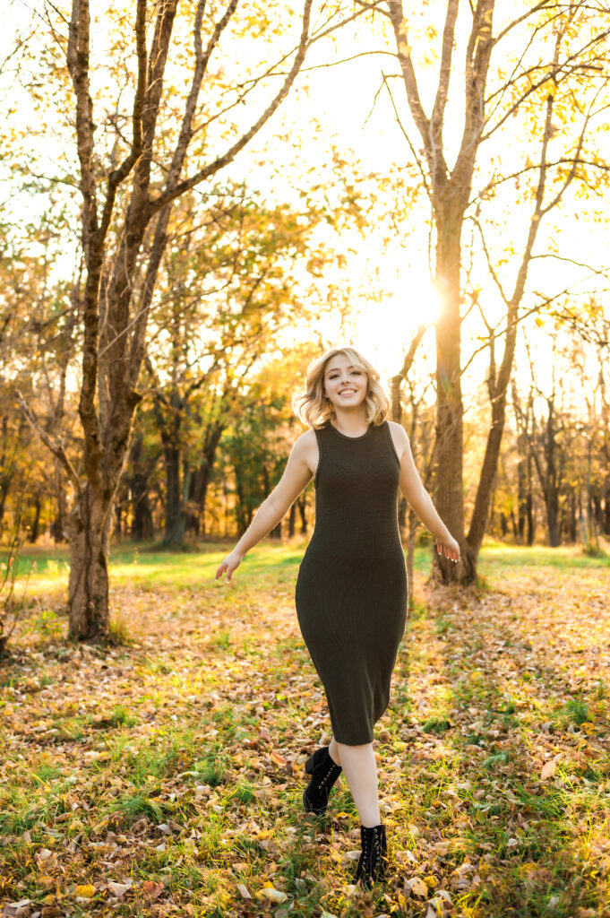 A high school teen wearing a long green sleeveless dress and boots smiles as she runs through trees during golden hour in the fall